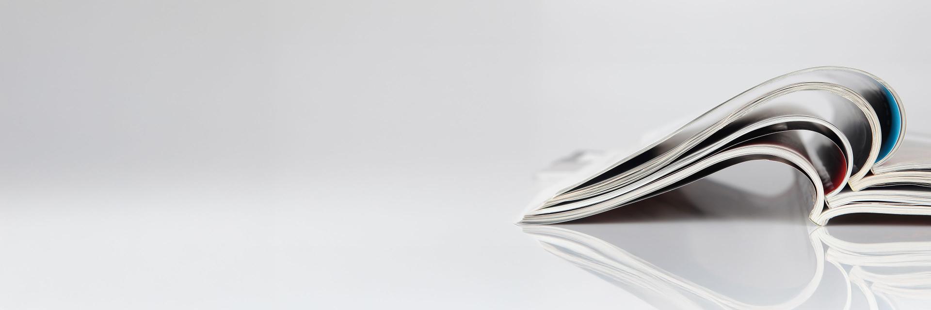 Close-up of two open magazines with pages curved into a wave-like shape, resting on a glossy white surface.