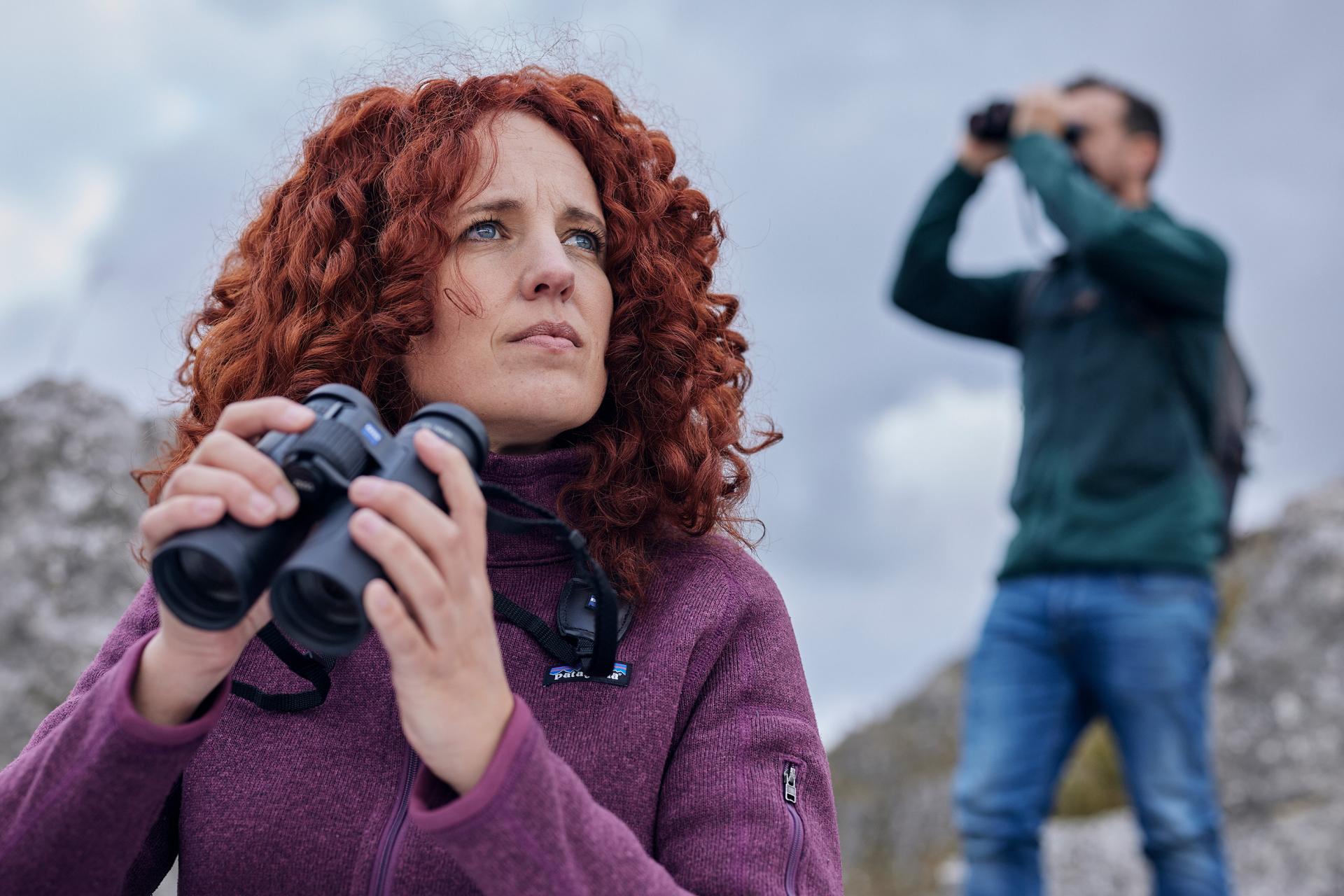 Two people outdoors using ZEISS binoculars, with a woman in the foreground and a man observing in the background.
