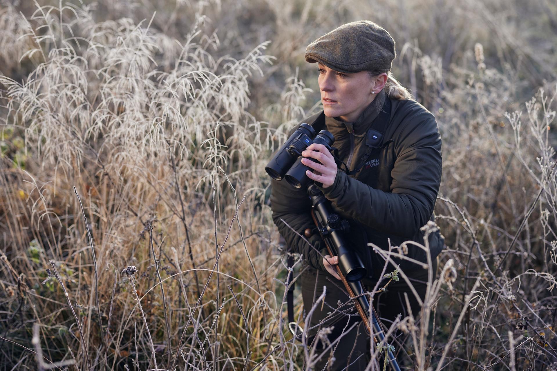 A person in outdoor gear holds a ZEISS binocular and a rifle while standing in a frosty field surrounded by tall grass.