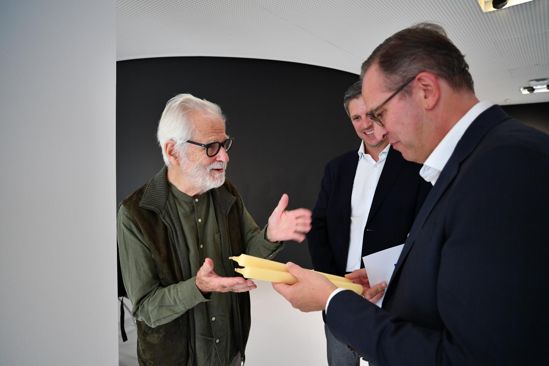 Three men in professional attire are engaged in a discussion while examining yellow candles.