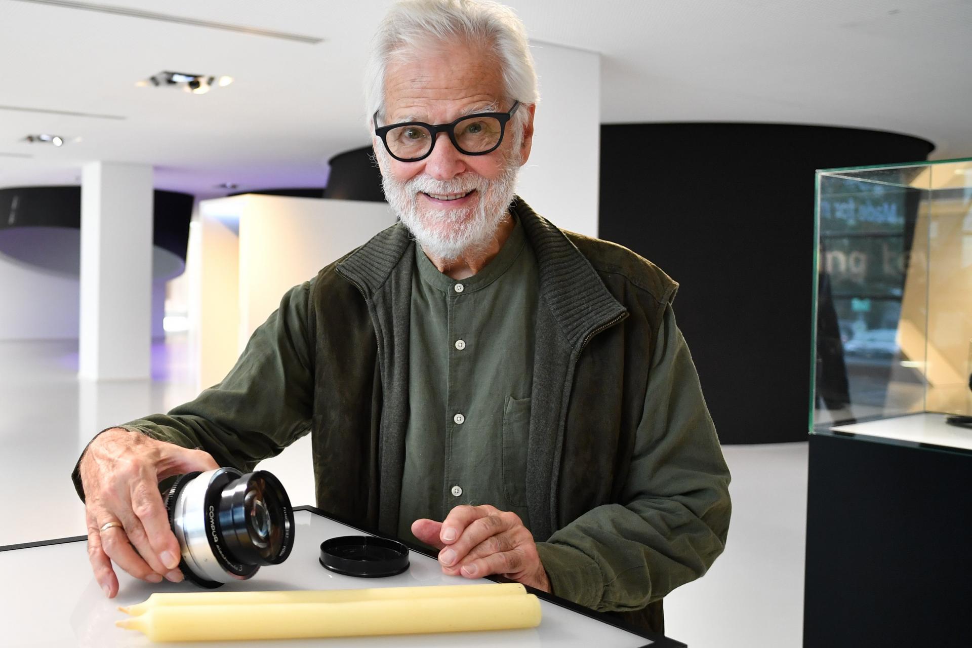 An older man with white hair and glasses is holding a ZEISS camera lens beside a lens cap and a long cylindrical object on a white table in a modern, brightly lit showroom.