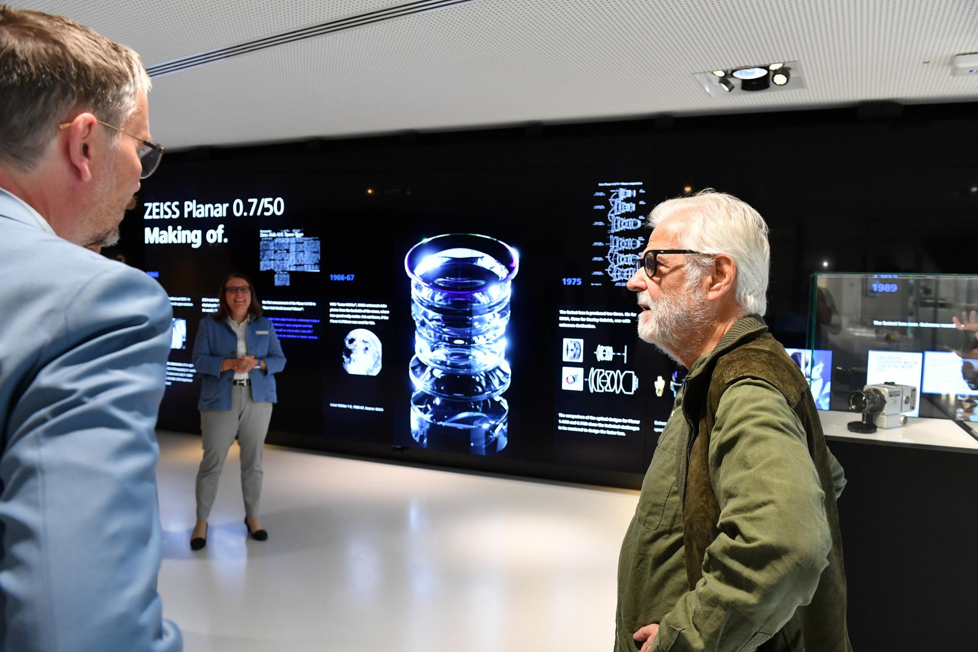 Three people are standing and conversing in front of a ZEISS display showcasing the making of the ZEISS Planar 0.7/50 lens with technical visuals and product information.