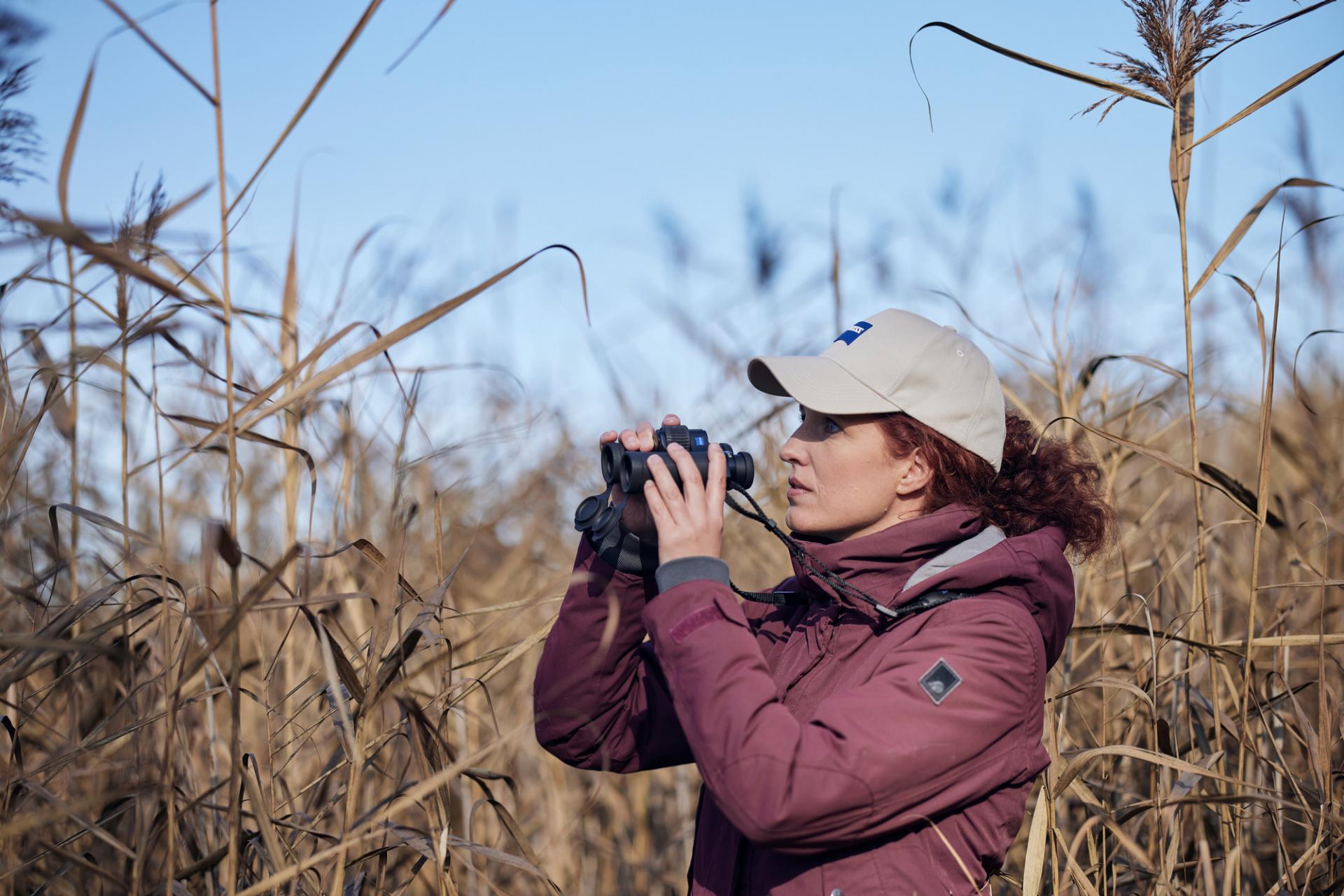A woman wearing a beige ZEISS cap and maroon jacket is observing through ZEISS binoculars in a field of tall, dry grass.