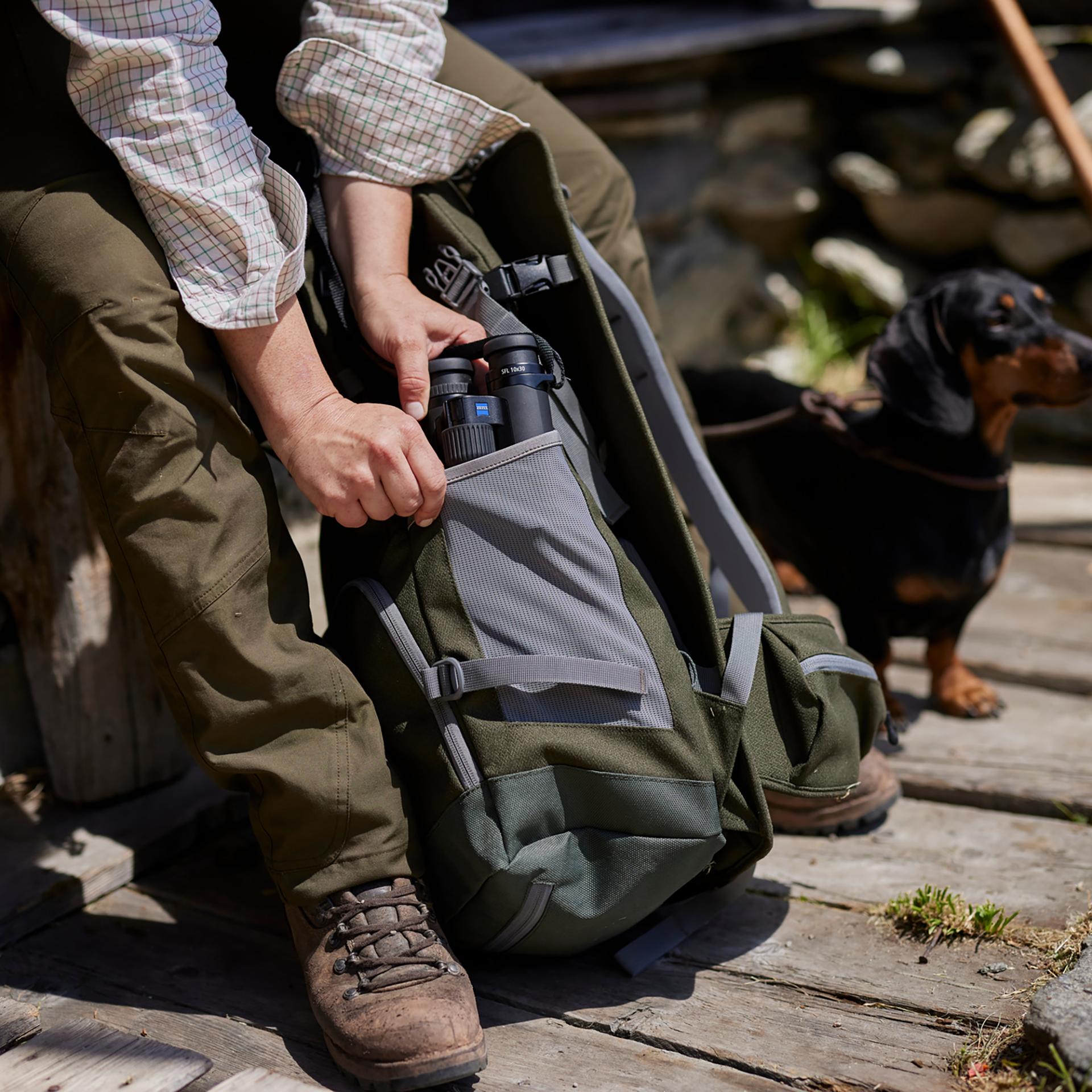 A person is placing ZEISS binoculars into the side pocket of a green backpack while sitting outdoors next to a small black and brown dog.