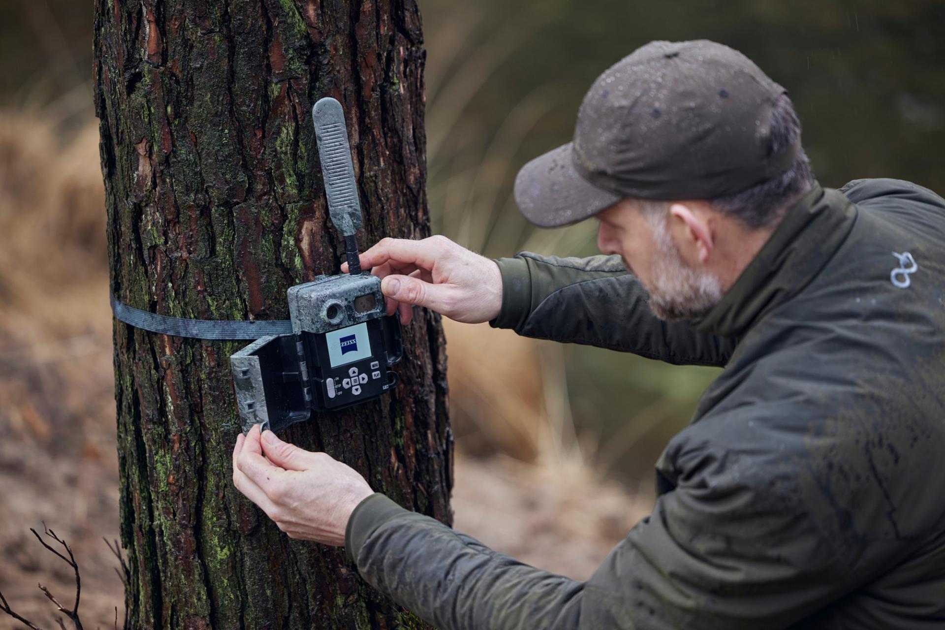 A man is mounting a ZEISS trail camera to a tree using a strap in an outdoor wooded area.