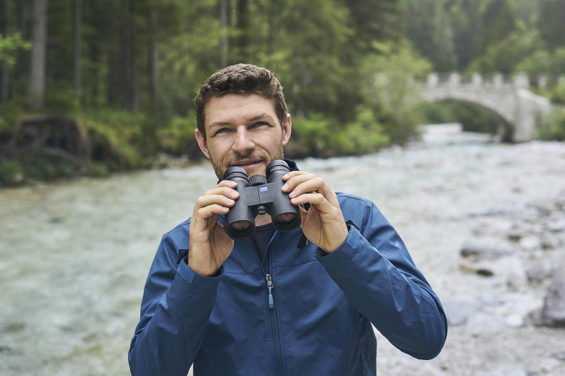A man in a blue jacket holds a pair of ZEISS binoculars while standing beside a river in a forested area with a stone bridge in the background.