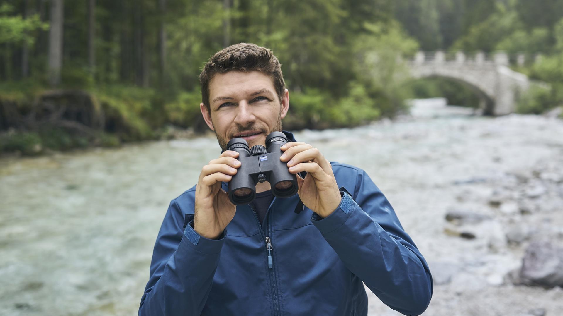 A man in a blue jacket holds a pair of ZEISS binoculars while standing beside a river in a forested area with a stone bridge in the background.