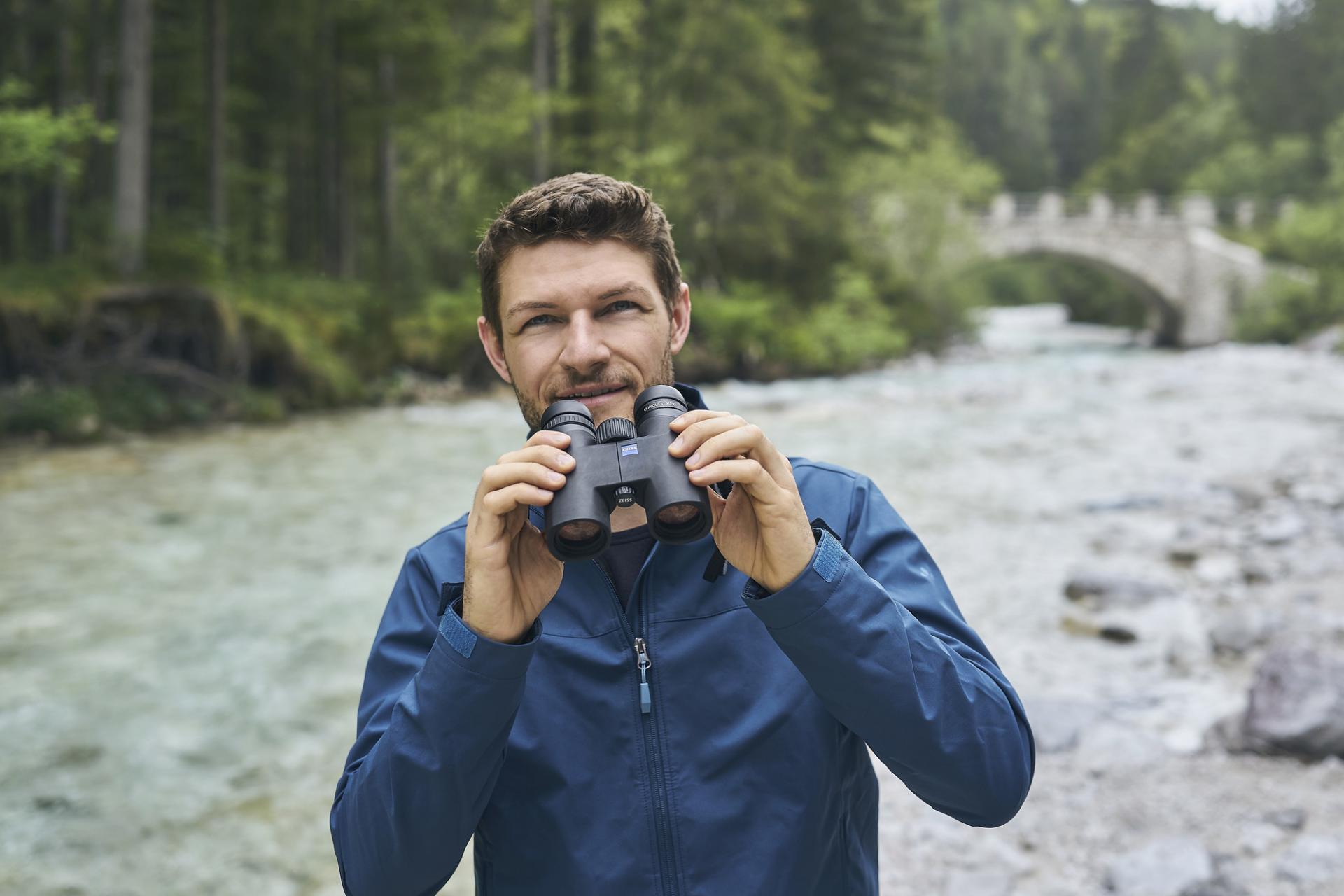 A man in a blue jacket holds a pair of ZEISS binoculars while standing beside a river in a forested area with a stone bridge in the background.
