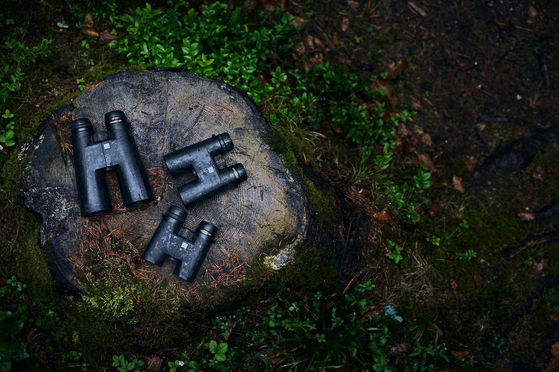 Three ZEISS binoculars are placed on a moss-covered tree stump in a forest setting.