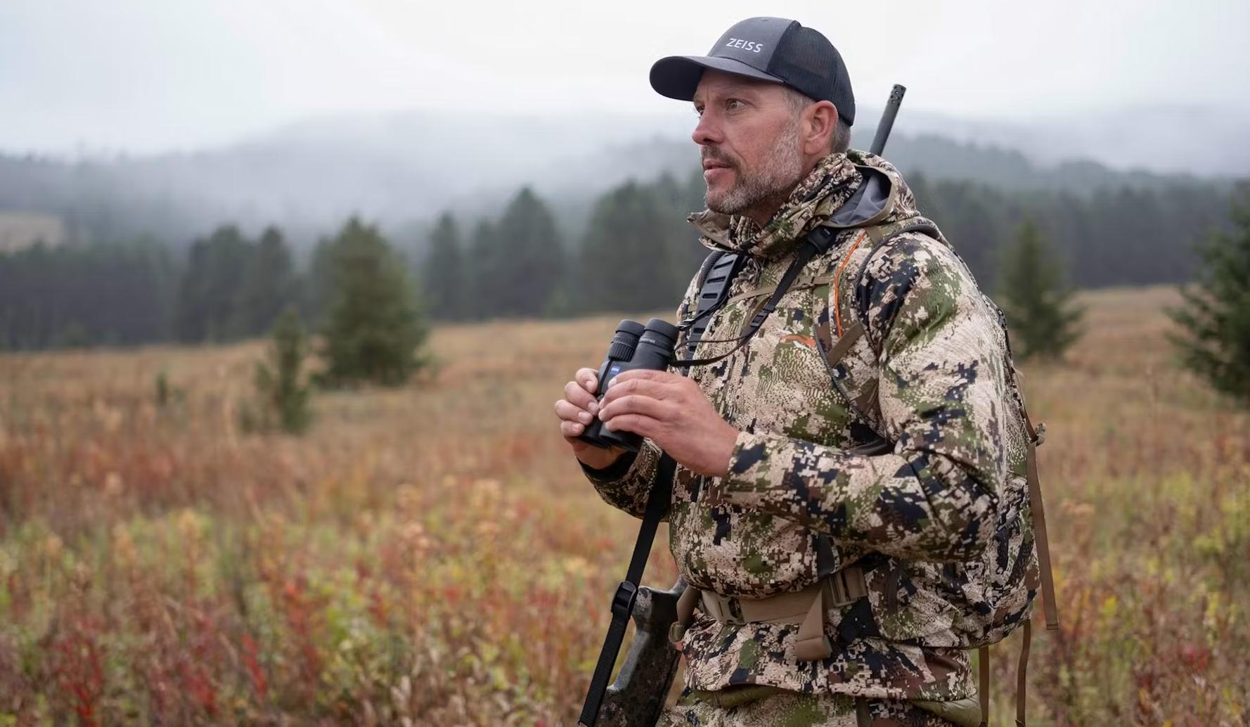 A man in camouflage clothing holding ZEISS binoculars stands in a misty, open field surrounded by trees and hills.