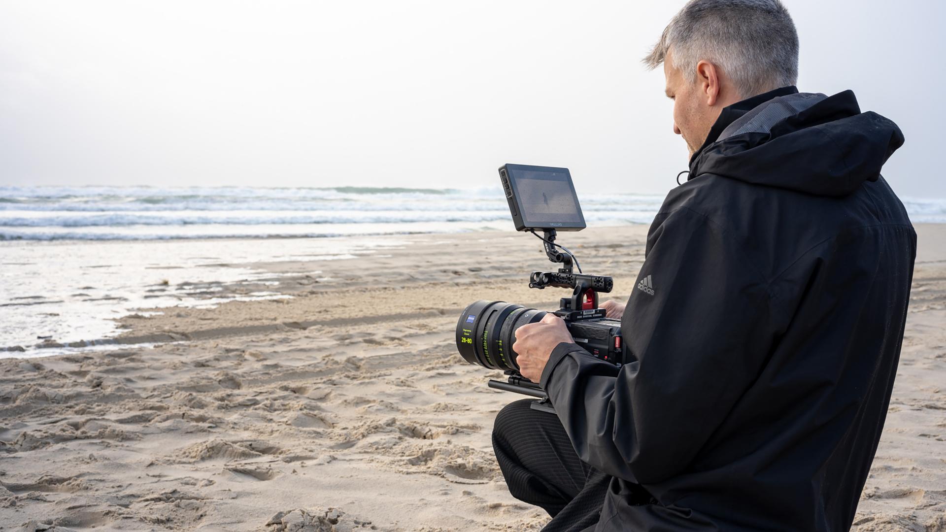A man kneels on a sandy beach while operating a professional video camera equipped with a ZEISS lens.