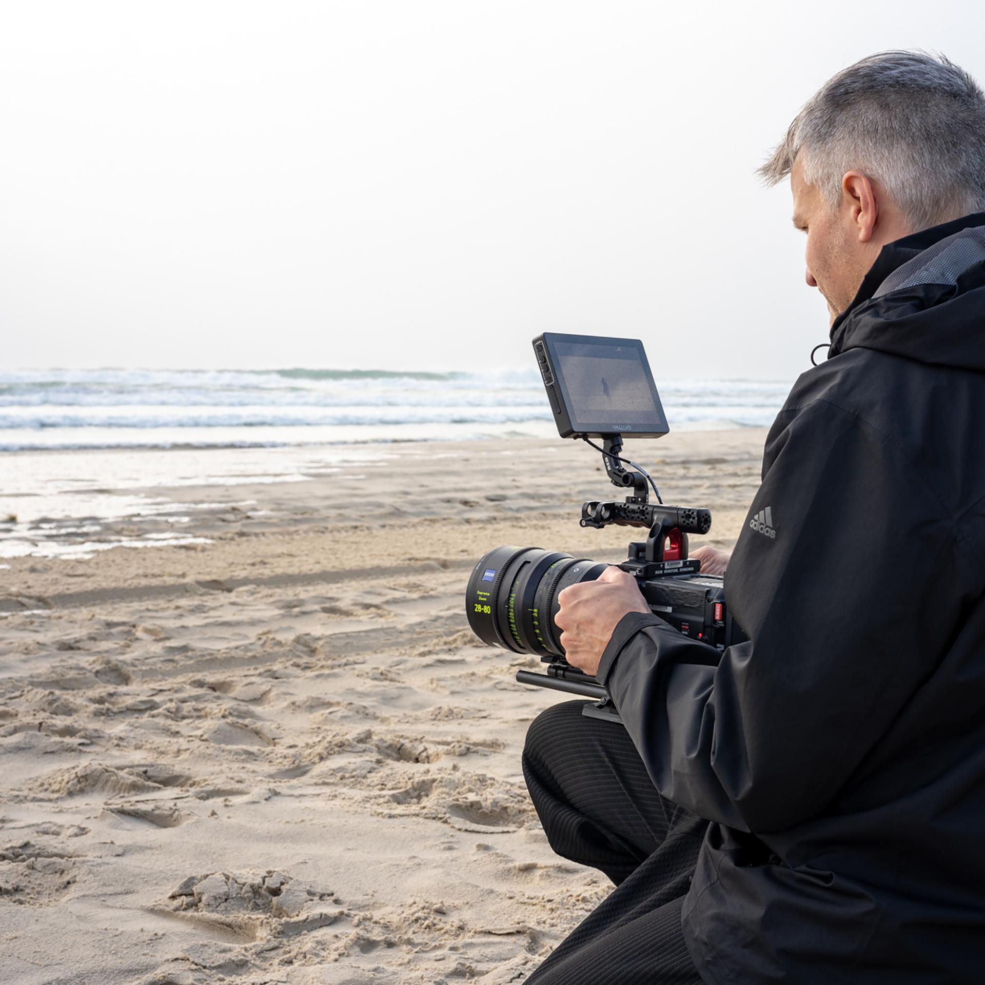 A man kneels on a sandy beach while operating a professional video camera equipped with a ZEISS lens.