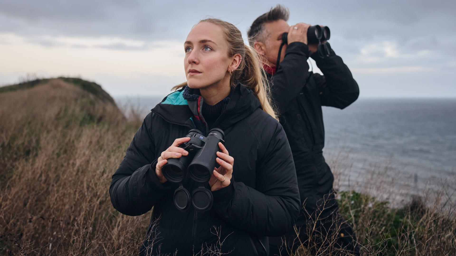 Two people standing on a coastal cliff holding ZEISS binoculars while observing the landscape.