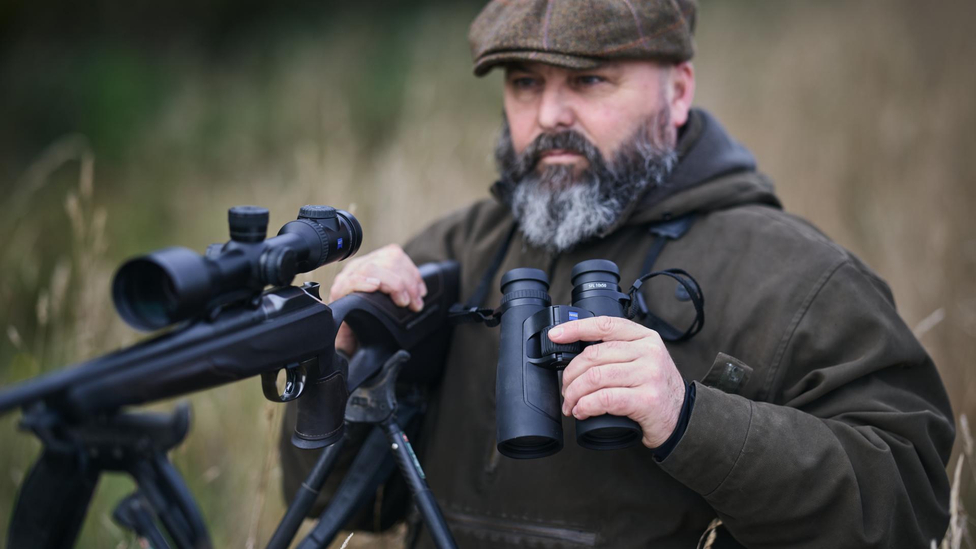 A bearded man in outdoor clothing stands in a field holding ZEISS binoculars and a rifle equipped with a ZEISS riflescope.