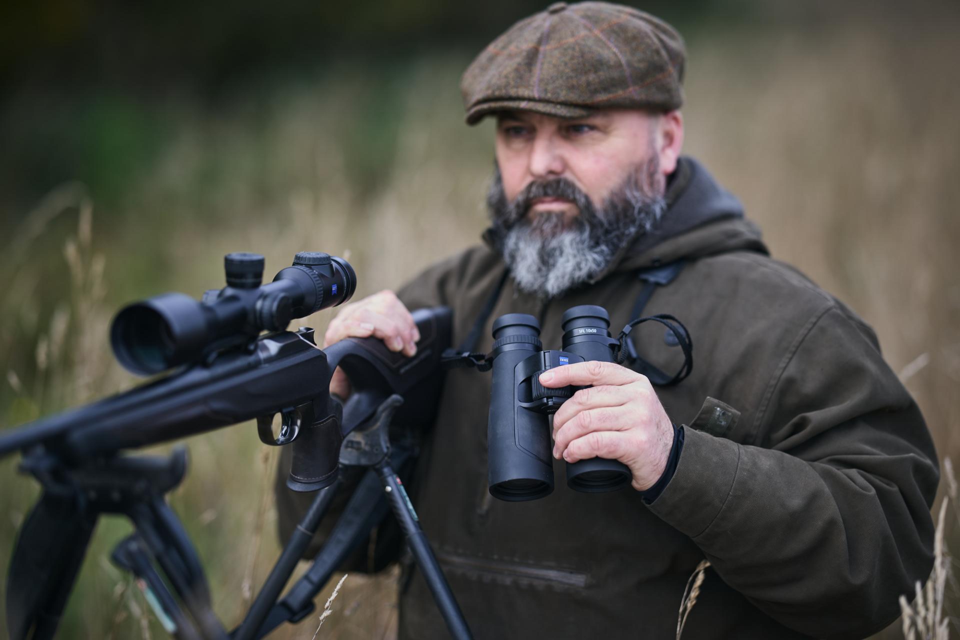Bearded man in outdoor clothing holding ZEISS binoculars and standing beside a rifle equipped with a ZEISS riflescope in a grassy field.