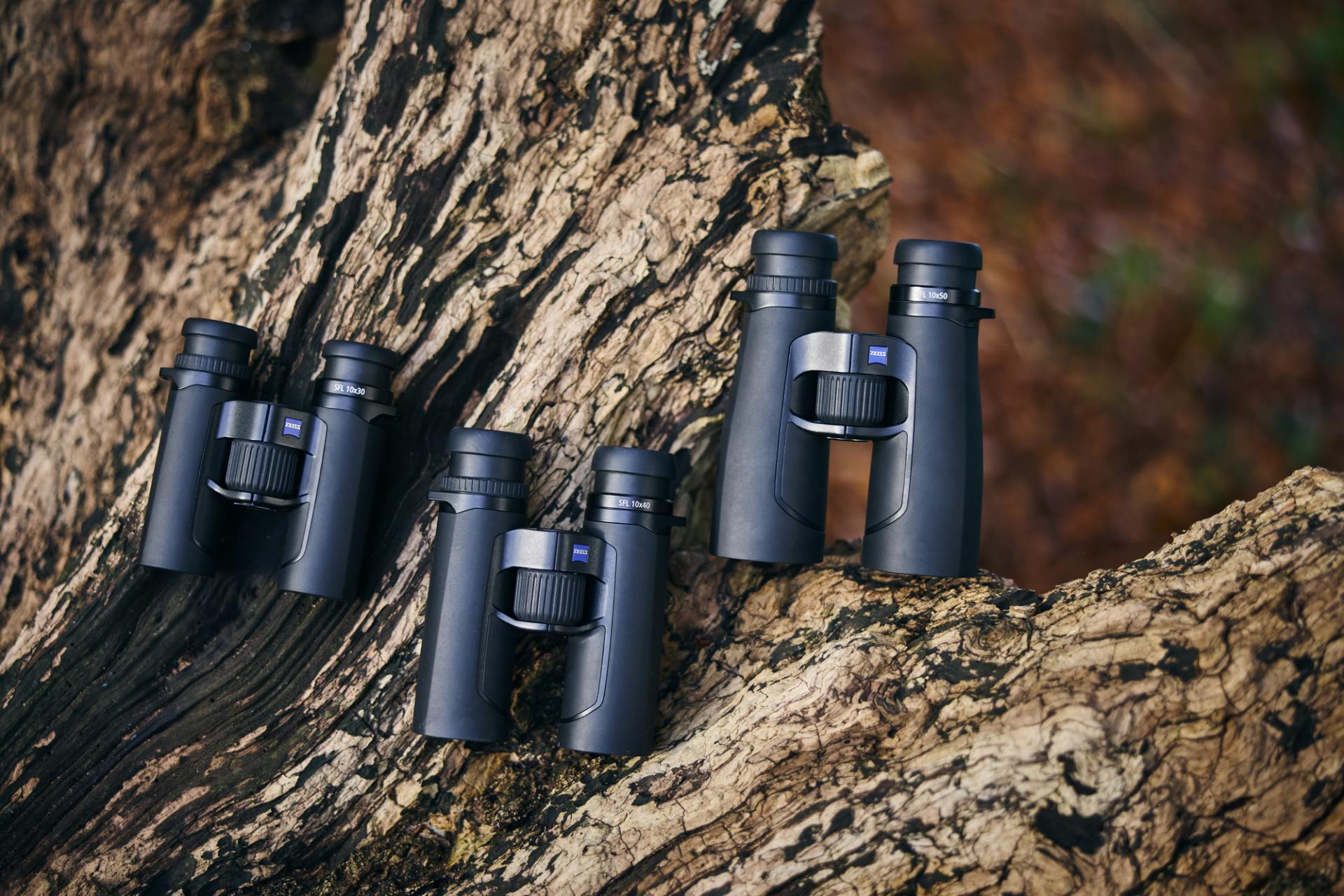 Three pairs of black ZEISS binoculars are displayed on the textured surface of a large tree trunk.