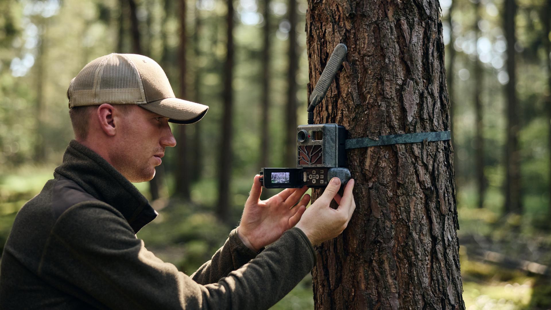 A man adjusts a ZEISS trail camera mounted on a tree in a sunlit forest.