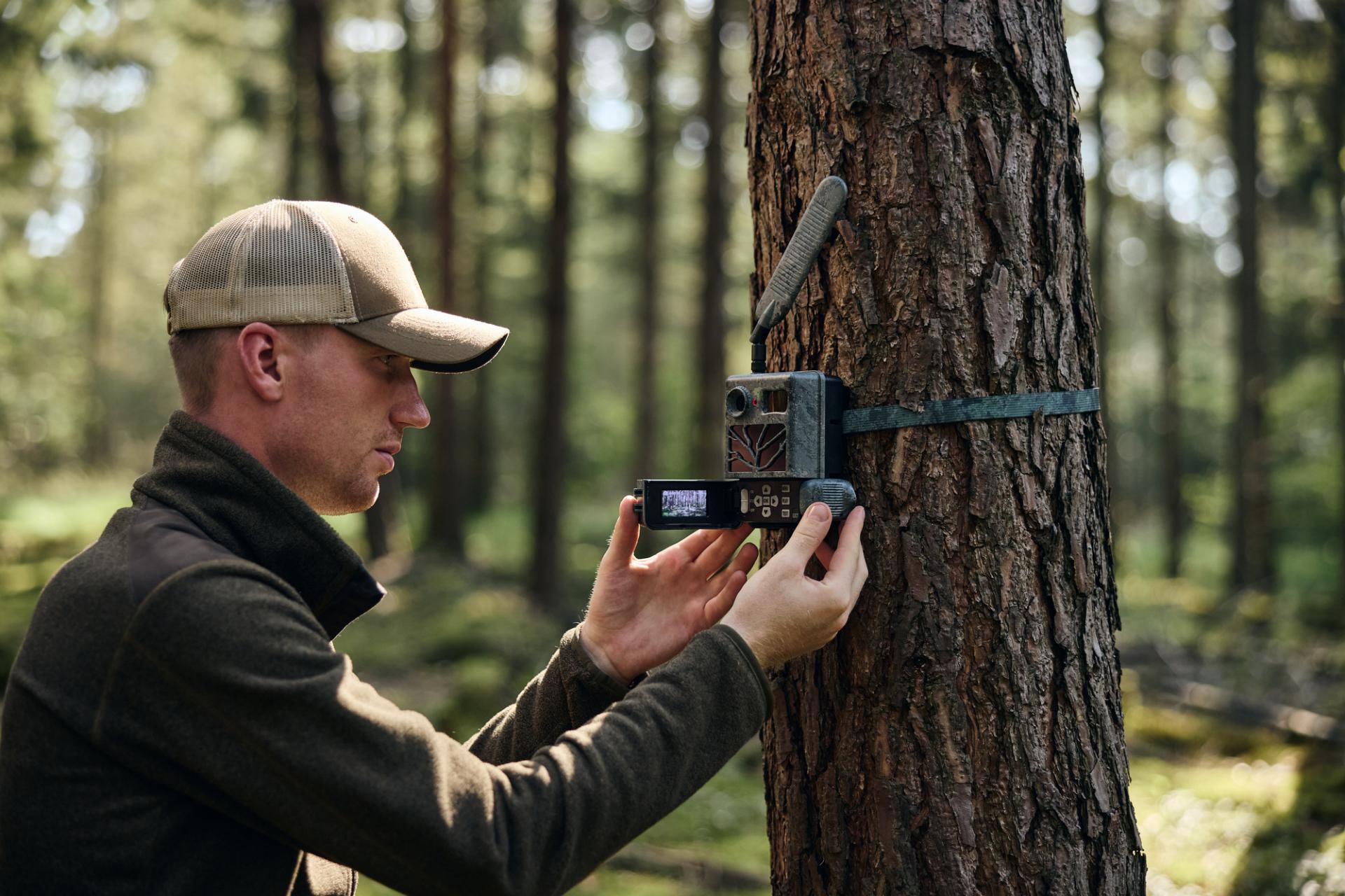 A man in outdoor clothing mounts a ZEISS trail camera to a tree in a forest.