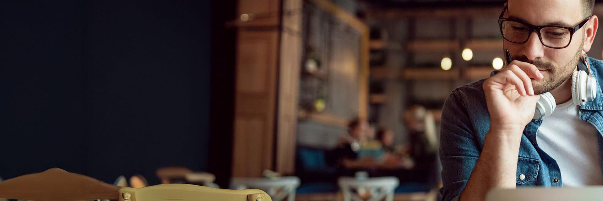 Man with glasses and headphones around his neck working on a laptop in a cozy, warmly lit café setting, wearing ZEISS lenses.