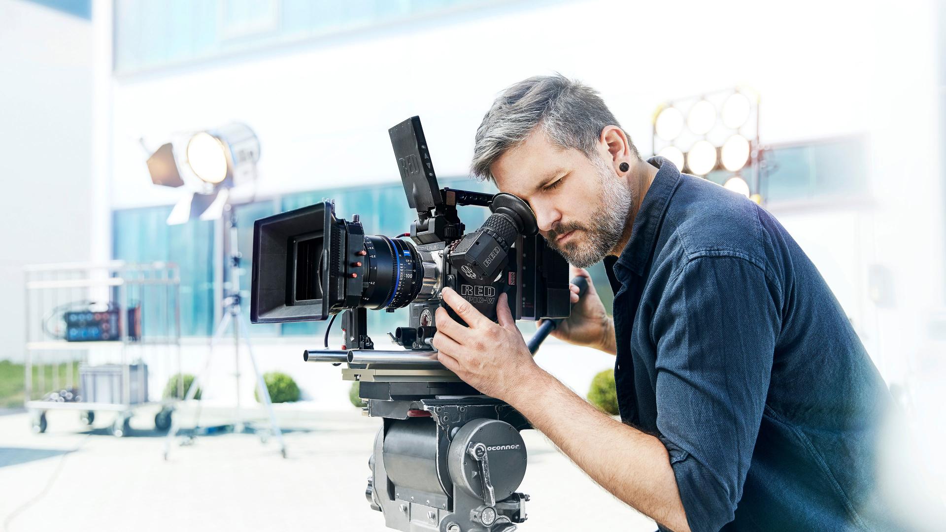 A man is operating a professional video camera equipped with a ZEISS lens on a film set with lighting equipment in the background.