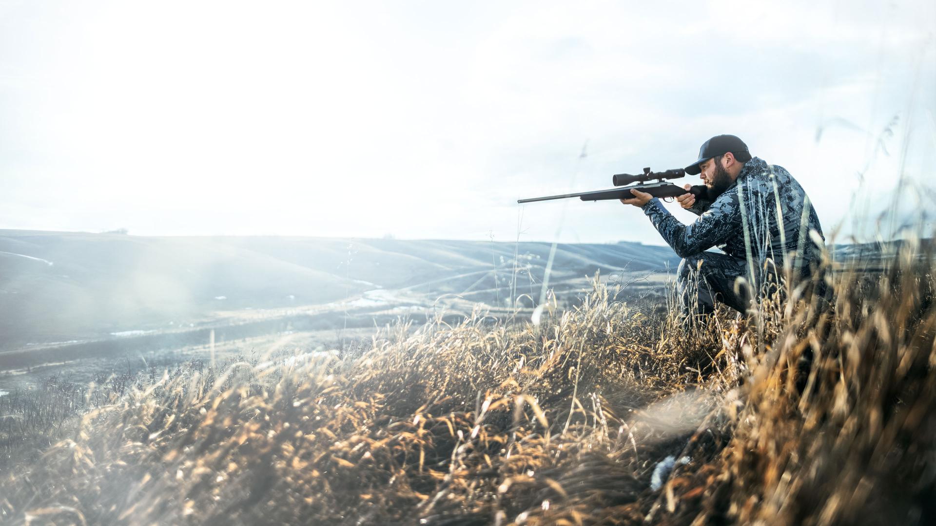 A man in camouflage gear aims a rifle equipped with a ZEISS scope while kneeling in a grassy, open landscape.