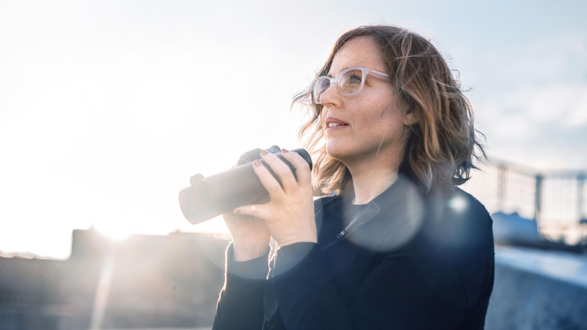 A woman wearing glasses is holding ZEISS binoculars while looking into the distance outdoors in bright sunlight.