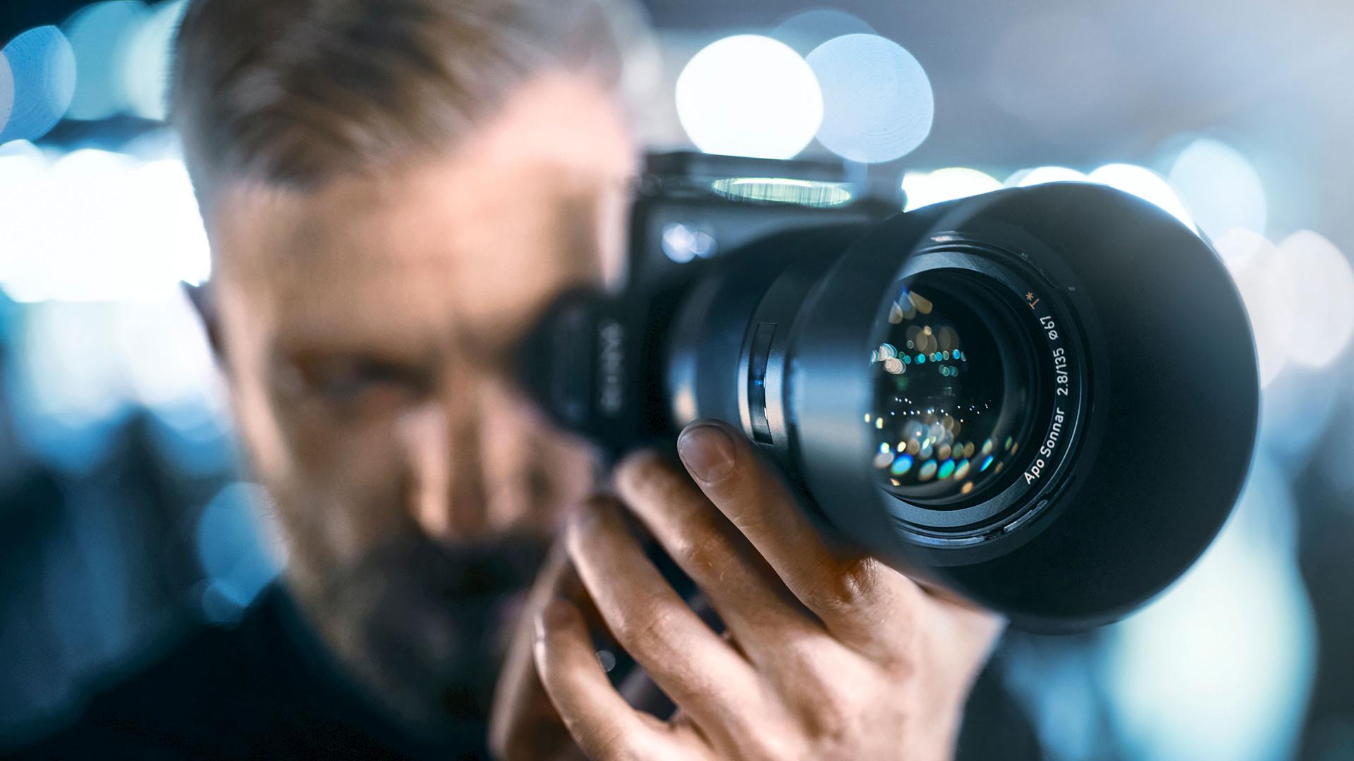 Close-up of a man holding a professional camera with a ZEISS lens aimed directly at the viewer.