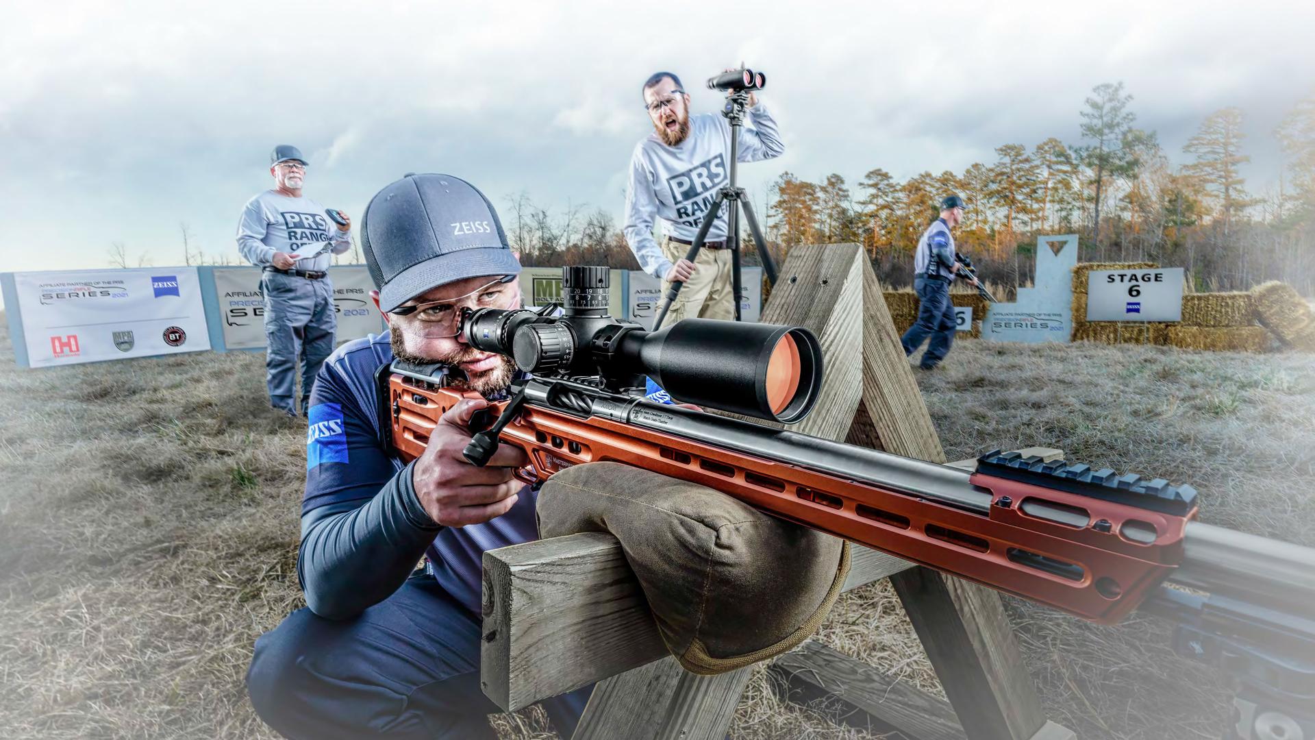 A competitive shooter aims through a precision rifle equipped with a ZEISS scope during an outdoor shooting event, with other participants and a stage sign visible in the background.