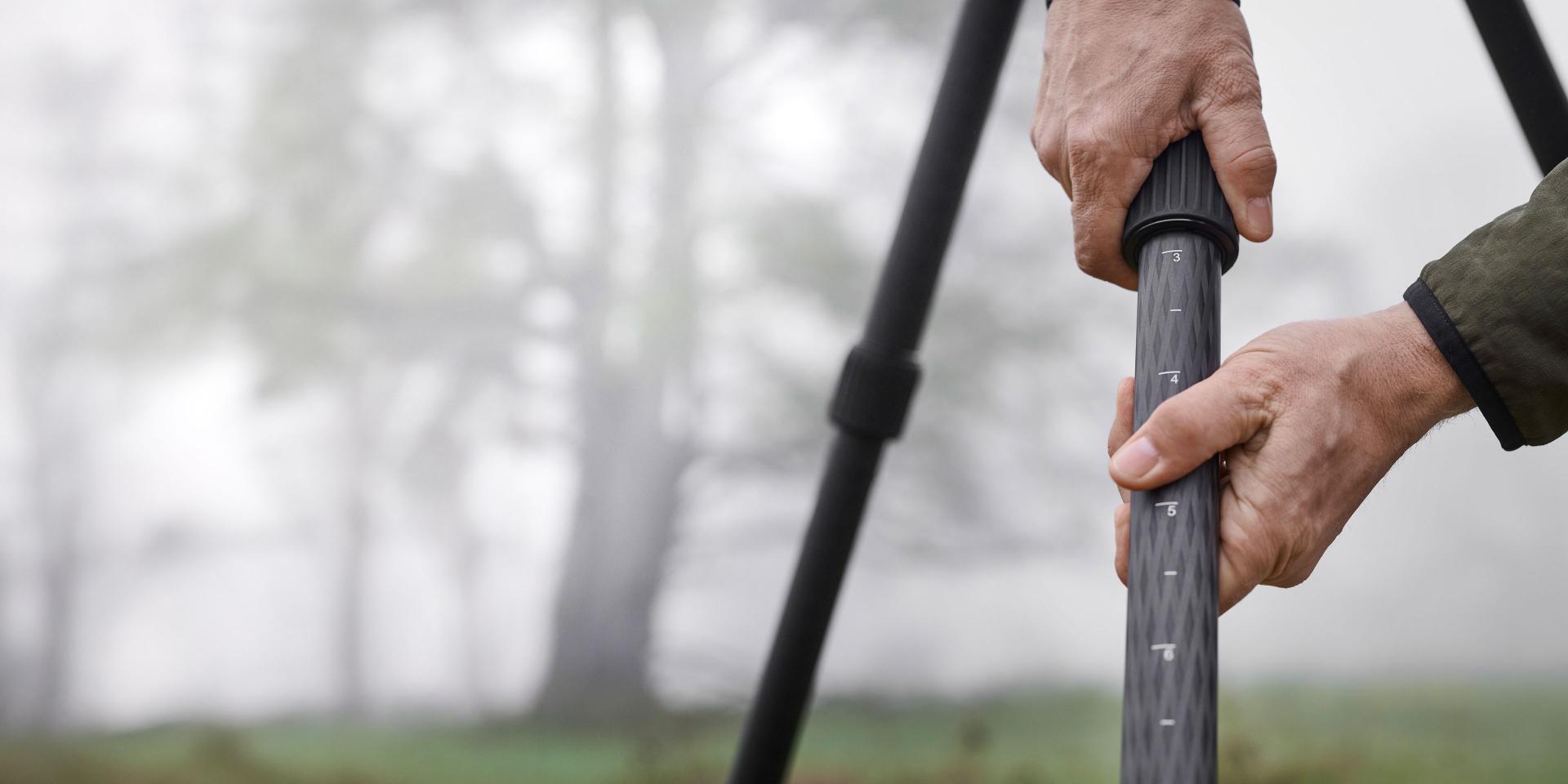 Close-up of a person adjusting the height of a tripod leg in an outdoor setting with foggy trees in the background.