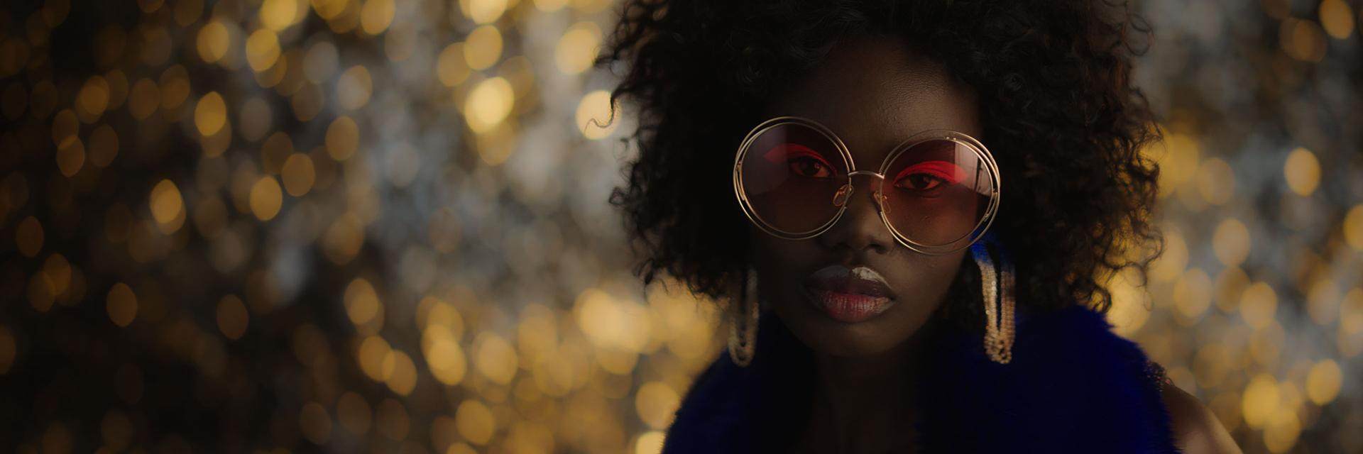 A woman wearing large, round sunglasses with red-tinted lenses and statement earrings poses in front of a golden, bokeh-lit background.