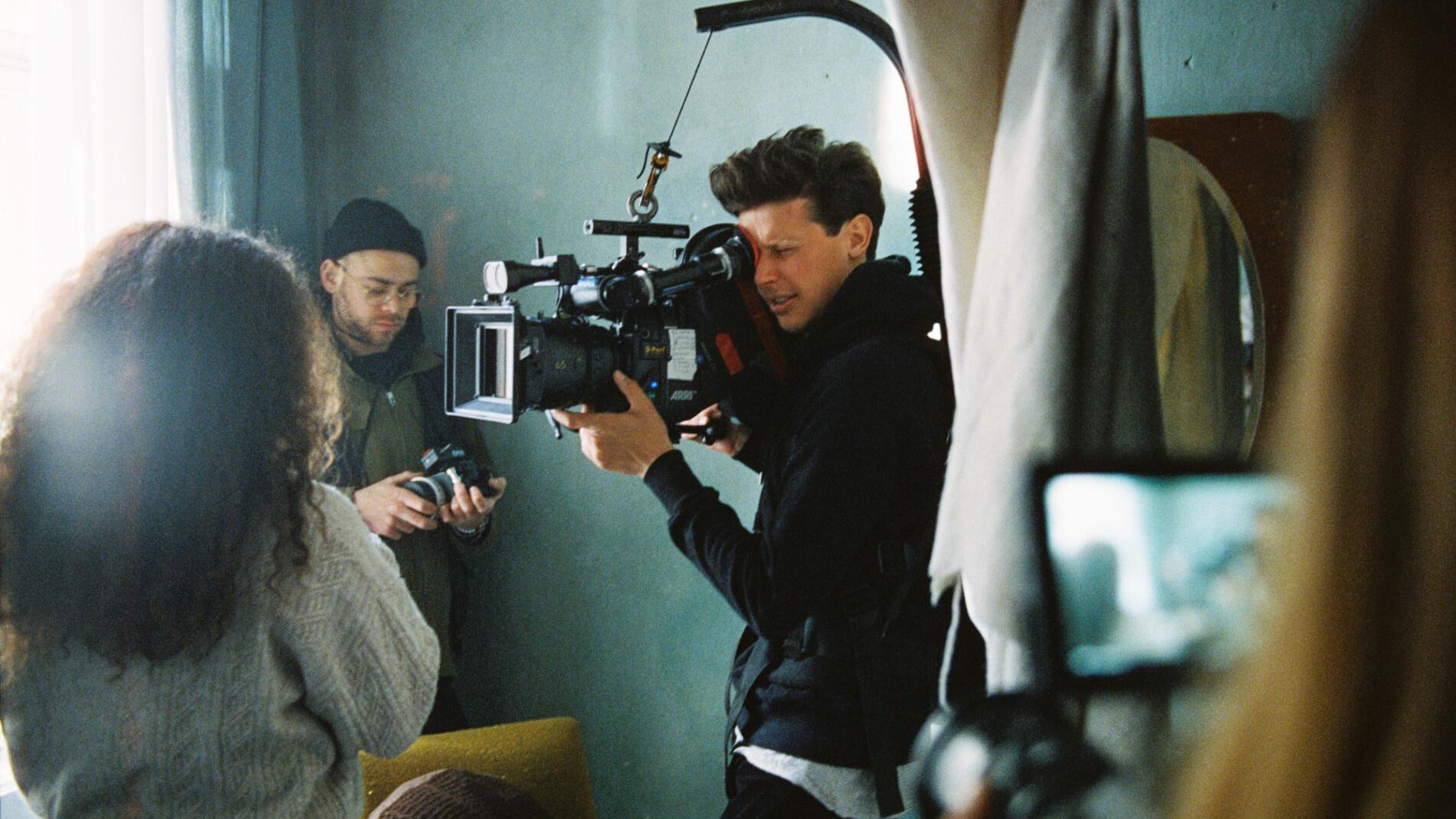 A cameraman operates a professional film camera equipped with a ZEISS lens during a video shoot with a small production crew.