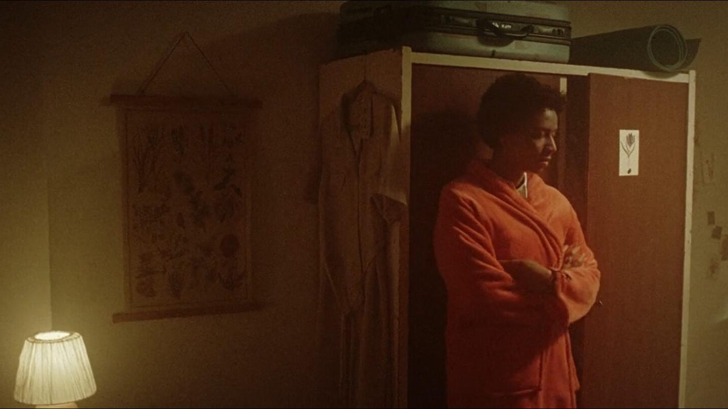A person in an orange robe stands with arms crossed next to a wardrobe in a dimly lit, warmly toned bedroom.