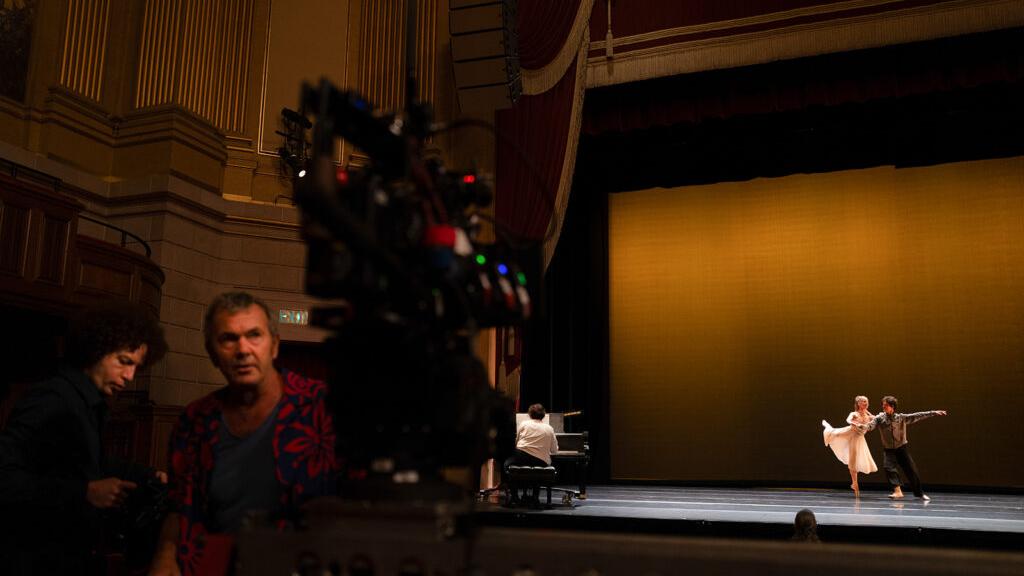A professional film camera setup captures a ballet performance on stage in a grand theater, with crew members working in the foreground.