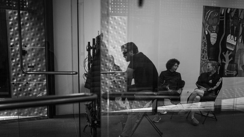 Three people are seated and engaged in conversation inside a modern, glass-walled room with contemporary artwork and technical equipment visible.