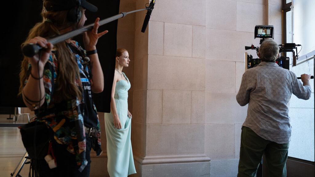 A film crew captures a woman in a light blue dress using professional camera equipment and a boom microphone in a well-lit indoor setting.