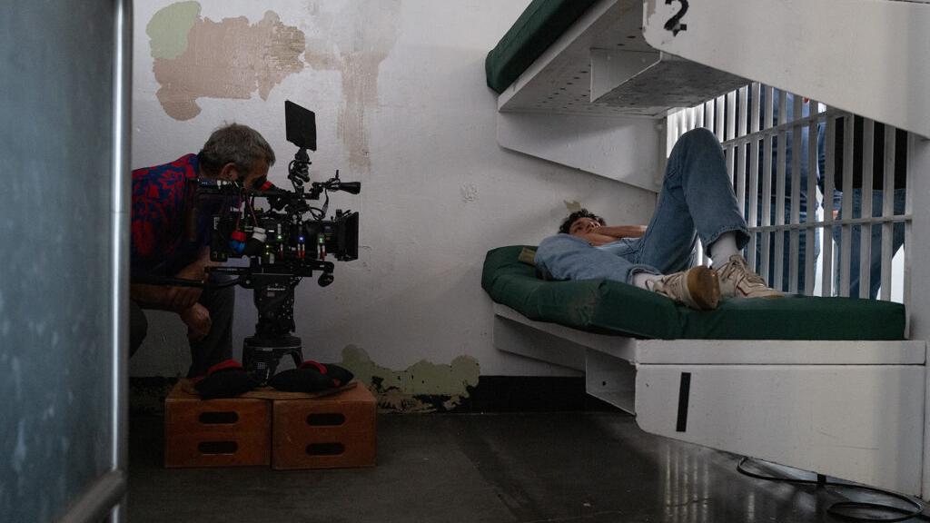 A camera operator films a scene inside a prison cell where an actor is lying on a bunk bed, using ZEISS cine lenses.