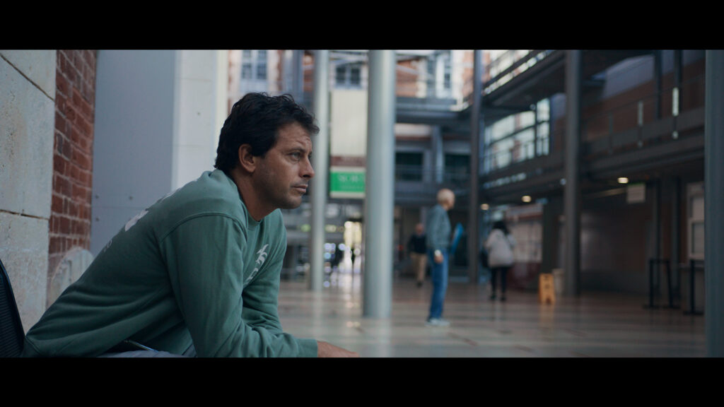 A man in a green sweatshirt sits on a bench in a modern indoor public space with people walking in the background, captured with clear depth and natural light using ZEISS optics.