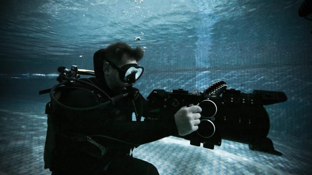 A diver in full scuba gear operates an underwater camera rig equipped with ZEISS lenses in a swimming pool.