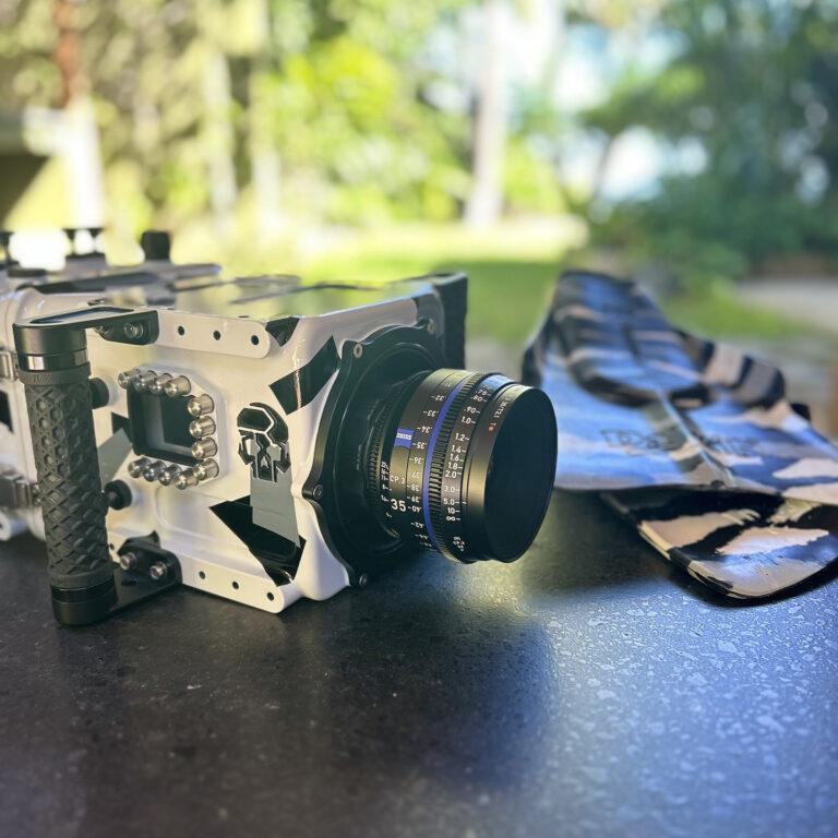 A professional cinema camera with a mounted ZEISS lens rests on a dark surface outdoors, with a soft focus background of greenery and sunlight.