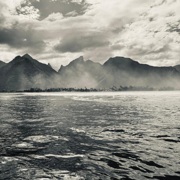 Black and white image of a calm ocean with gentle waves in the foreground and a misty mountain range under a partly cloudy sky in the background.