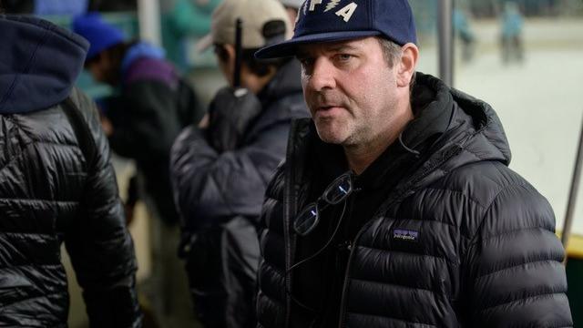 A man wearing a black puffer jacket and a dark blue cap stands indoors near an ice rink, with several other people in the background.