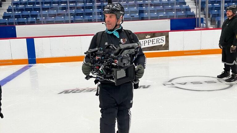 A camera operator wearing ice skates and professional gear films on an ice hockey rink, holding a stabilized camera rig possibly fitted with ZEISS lenses.