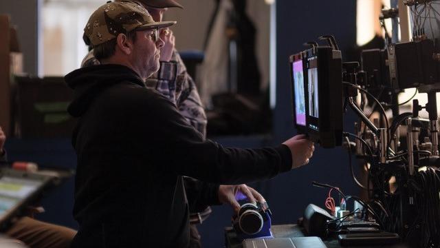Two technicians operate professional video monitors and equipment on a film set, with one holding a ZEISS lens.