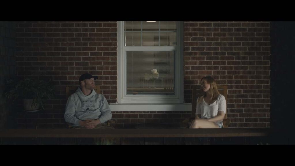 A man and a woman sit apart on a wooden bench outside a brick building at night, with a window and a potted plant between them.