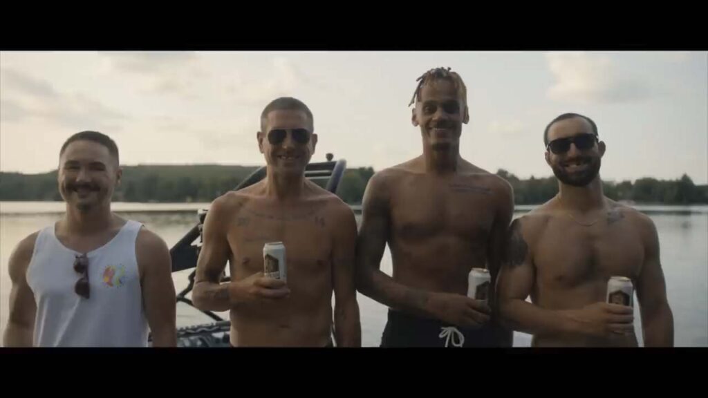 Four smiling men, three shirtless and one in a tank top, stand by a lakeside holding canned drinks with a boat and trees in the background.