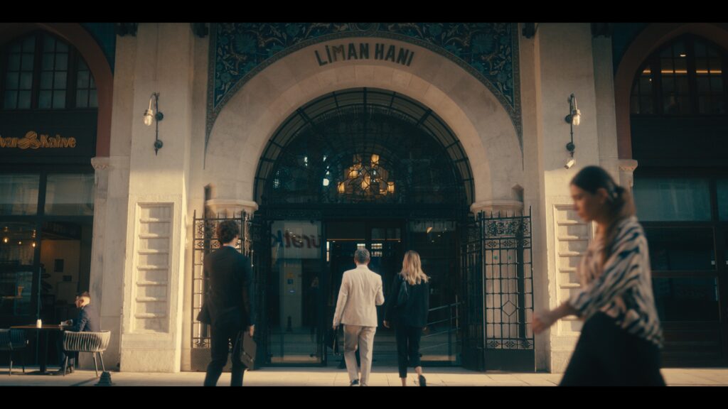 Several people walk toward the entrance of a grand building labeled "LIMAN HANI," featuring ornate architecture and a hanging chandelier.