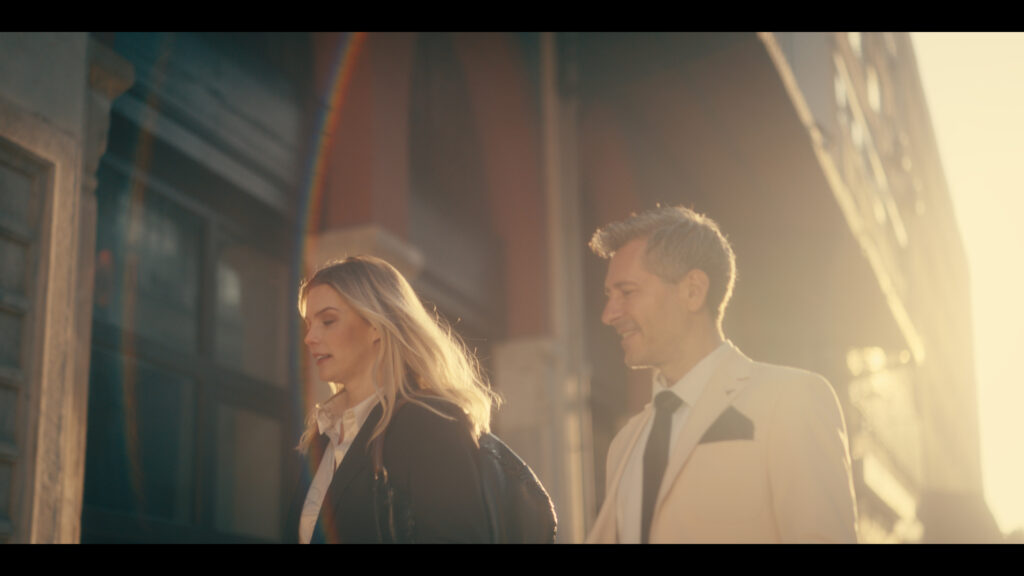A professionally dressed woman and man walk together outside a sunlit building, captured with a warm, high-quality image typical of ZEISS optics.