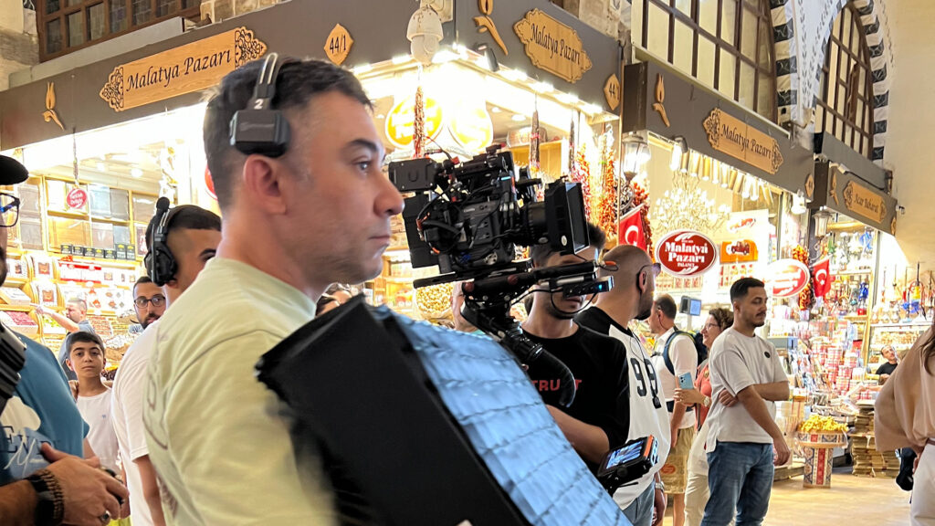 A film crew equipped with professional camera gear, including a ZEISS lens, shoots in a bustling indoor market filled with colorful storefronts and shoppers.
