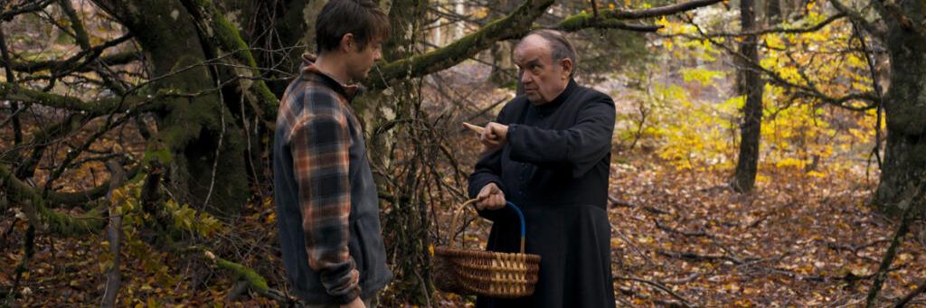 A man in a plaid shirt stands in a forest opposite an older man in a black coat holding a woven basket and pointing, surrounded by autumn trees.