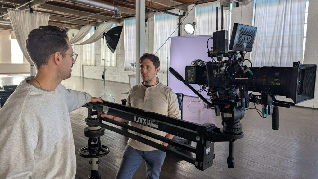 Two men stand next to a professional film camera mounted on a ZEISS camera rig inside a well-lit studio with large windows and photography equipment.